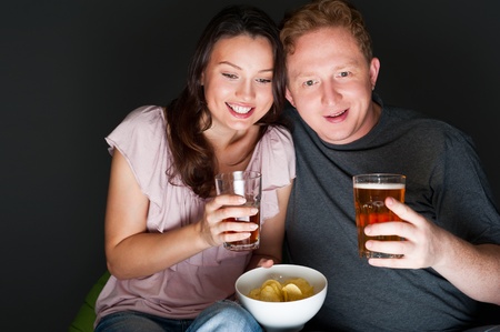 Happy couple sitting together watching something intresting on television drinking and eating - Grey backgroundの写真素材