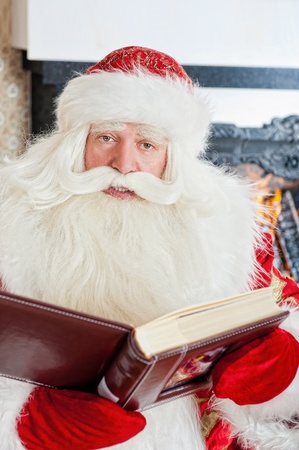 Santa sitting at the Christmas tree, fireplace and reading a bookの写真素材