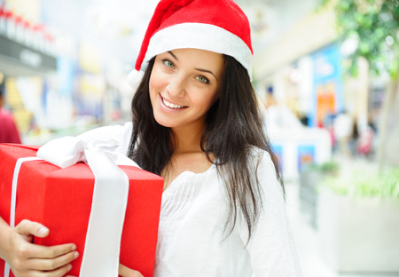 Portrait of young pretty woman wearing Santa Claus helper hat standing inside shopping mall smiling and holding christmas giftの写真素材