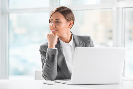 Thoughtful business woman holding chin while using laptop at workの写真素材