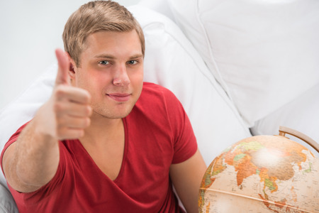 Portrait of a handsome young man sitting at home with globe and thumbs upの写真素材