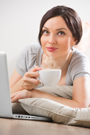 A young woman lying on the floor in front of her laptop with cup of tea or coffeeの写真素材