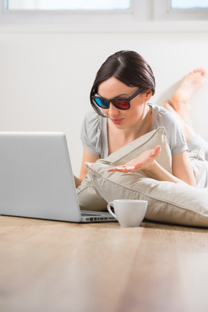 A young woman lying on the floor in front of her laptop with cup of tea or coffee wearing 3d glasses and watching movie with expressive emotionsの写真素材