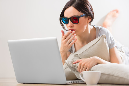 A young woman lying on the floor in front of her laptop with cup of tea or coffee wearing 3d glasses and watching movie with expressive emotionsの写真素材