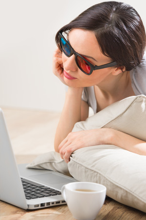 A young woman lying on the floor in front of her laptop with cup of tea or coffee wearing 3d glasses and watching romantic or comedy movie の写真素材