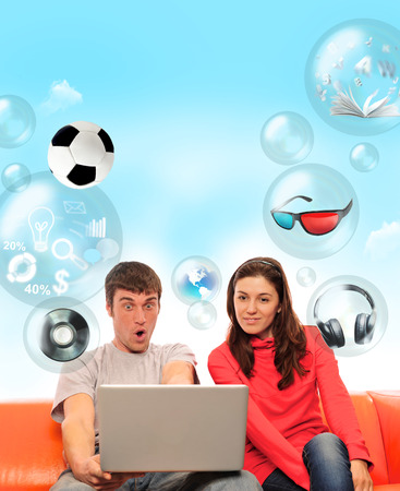 Adult couple sitting on sofa and having fun using their laptop. Different objects flying around themの写真素材