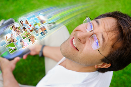 Adult man sitting on grass in park and using social media to look through new photo of his friendsの写真素材