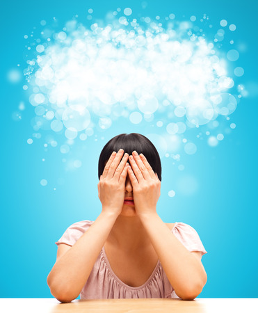 Young woman sitting against blue background with eyes closed by her hands. Blank cloud balloon with copyspace overheadの写真素材