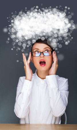 Beautiful European young solid businesswoman sitting with glasses against grey background and thinking about something. Blank cloud balloon with copyspace overheadの写真素材