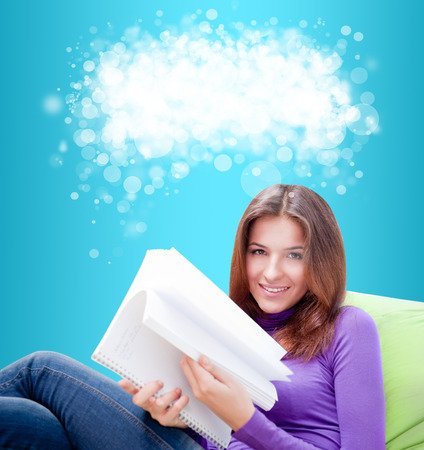 Young adult woman writing in her copybook while sitting on a large beanbag against blue background. Blank cloud with copyspace overheadの写真素材