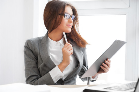Beautiful business woman looking at papers she holding in her arms while working on computer at her officeの写真素材