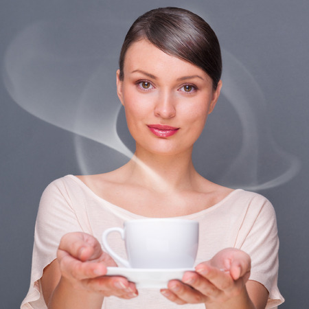 Portrait of young fashionable woman giving hot coffee or tea beverage to camera against grey backgroundの写真素材