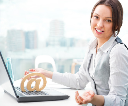 Portrait of a cheerful Business woman sitting on her desk and smiling. At symbol on her laptopの写真素材