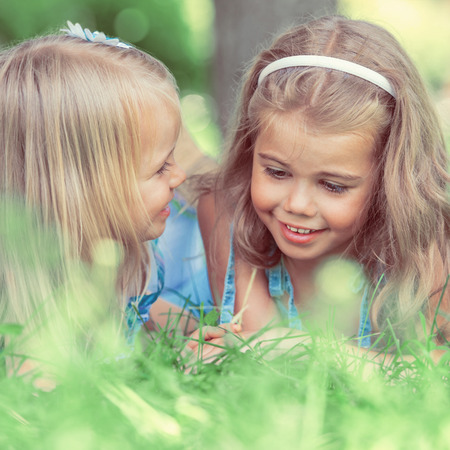 Two little sisters having fun in summer park while laying on lawn. Retro filter photoの写真素材