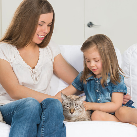 Mother and daughter sitting on sofa and cuddling catの写真素材