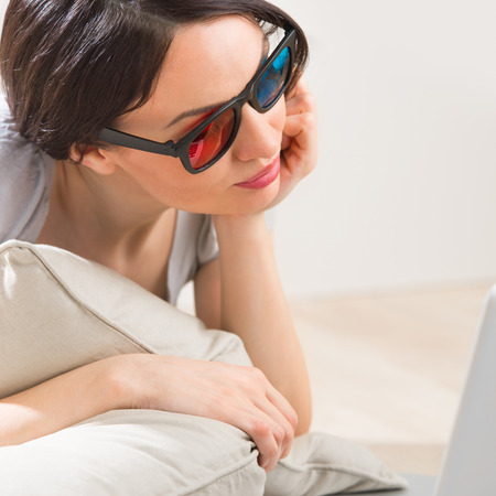 A young woman lying on the floor in front of her laptop with cup of tea or coffee wearing 3d glasses...