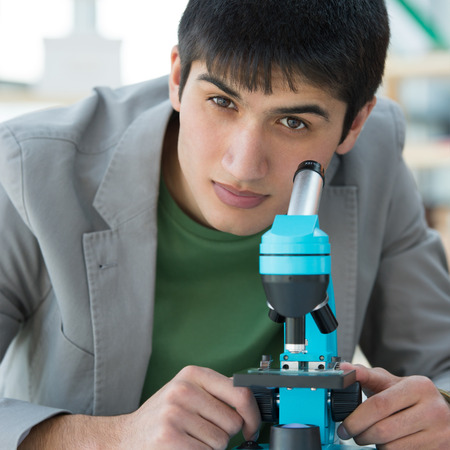Male student in laboratory looking at camera while working with microscope and making notes