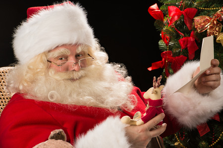 Santa Claus sitting at home at comfortable armchair holding envelope and reading children's letters and wishes and choosing toys from big sack near him. の写真素材