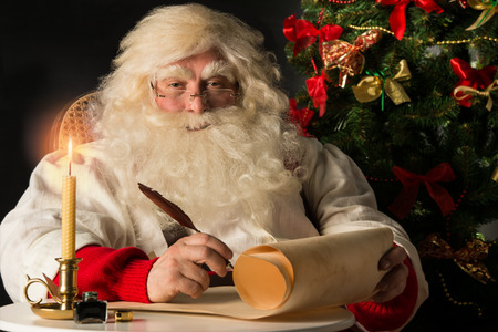 Santa Claus sitting at home and writing on old paper roll to do list with quill pen and ink at night with candle light. Authentic vintage style portrait.の写真素材
