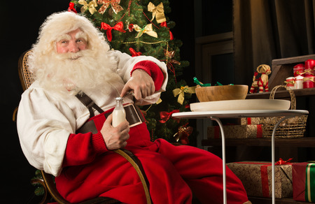 Santa Claus eating cookies with milk sitting near Christmas tree. Opening milk bottleの写真素材