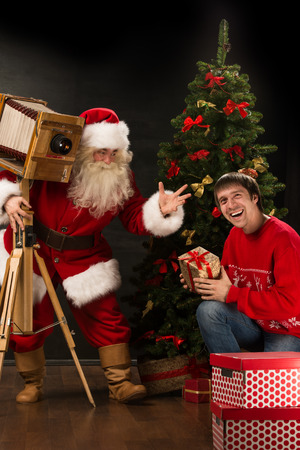 Santa Claus taking picture of cheerful man with old wooden camera at home near Christmas treeの写真素材