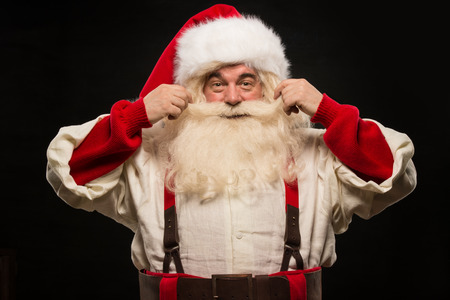 Photo of happy Santa Claus touching mustache and looking at camera on dark backgroundの写真素材