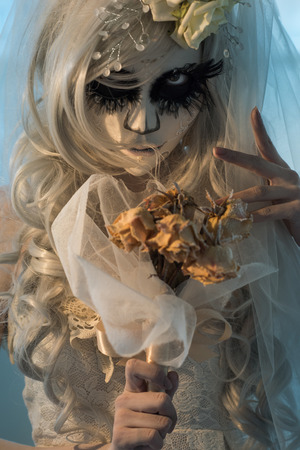 Halloween witch. Beautiful bride woman wearing santa muerte mask and wedding dress holding dead bouquet of rosesの写真素材
