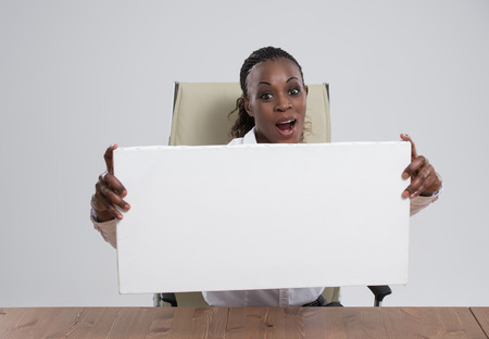 Smiling African Business woman portrait sitting at workplace with blank white board on gray isolated background Female model with long hair.の写真素材