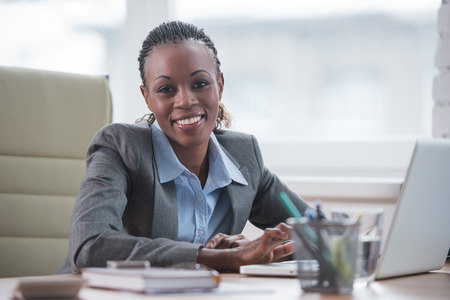 Pretty cheerful african businesswoman working with laptop in office and looking at cameraの写真素材
