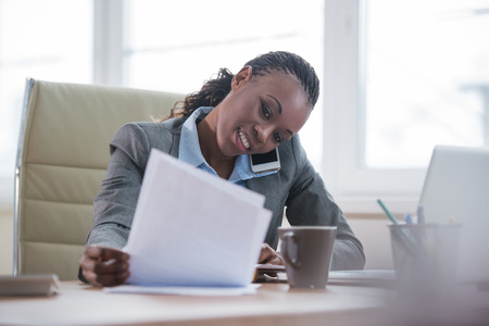 Portrait of businesswoman working with laptop writes on a document and calling phone at her officeの写真素材