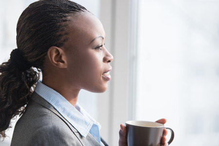 Pretty african businesswoman standing near window and drinking coffee or tea while resting and thinking about somethingの写真素材