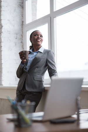 Happy african business woman enjoying the coffee break at her office and looking at windowの写真素材