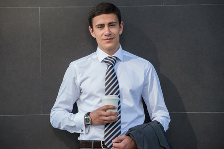 Businessman drinking coffee leaning on wall of modern office building. Urban professional smiling happy wearing white shirt holding disposable coffee cup. Handsome male model in his twenties.の写真素材