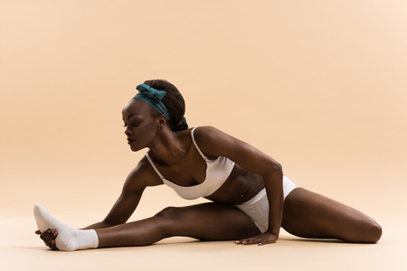 Portrait of young african woman doing stretching exercise on beige backgroundの写真素材