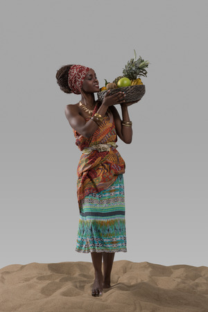 Attractive young african woman carrying fruits on sand on gray studio backgroundの写真素材