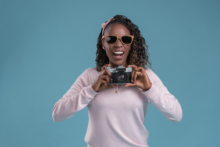 Beautiful young african woman using camera and photographing on blue backgroundの写真素材