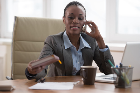 Business woman calling phone and writing something in her schedule book and papers at office workplaceの写真素材