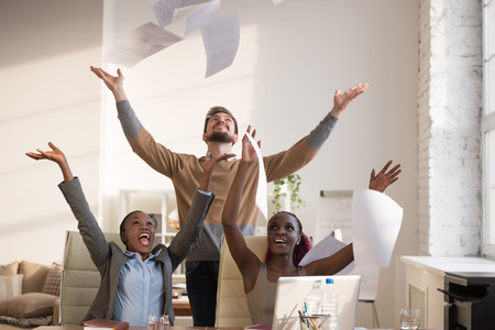 Business people excited happy smile, throwing up papers, documents fly in air, businesspeople sitting at office desk hold hands arms up, success team conceptの写真素材