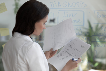 Female medical doctor working at clinic office. Writing on glass whiteboard symptoms and test results of her patient to diagnose diseaseの写真素材
