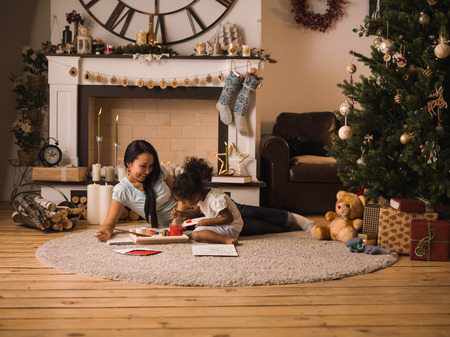 Mixed race mother and daughter playing and painting near Christmas tree at homeの写真素材