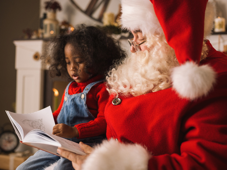 Santa Claus reading a book with cute african child near the fireplace and Christmas tree at homeの写真素材