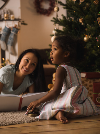 Mixed race mother and daughter reading book near Christmas tree at homeの写真素材