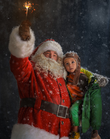 Photo of Santa Claus standing outdoors under snow with Child and burning sparklerの写真素材