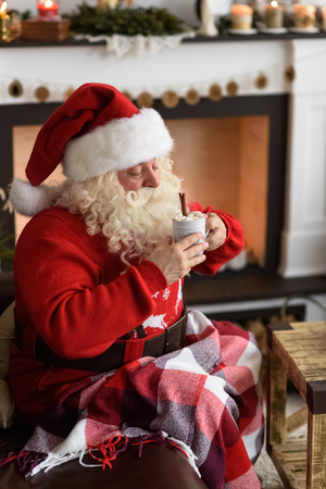 Santa Claus Drinking Coffee with Marshmallow near Fireplace at Homeの写真素材