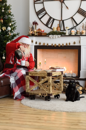 Santa Claus Drinking Coffee with Marshmallow near Fireplace at Homeの写真素材
