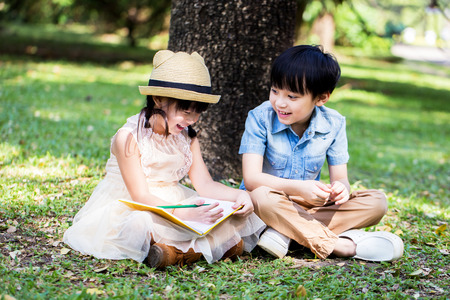 Little asian girl use pencil writting on notebook for writing book with smiling face in the park and her brother looking to herの写真素材