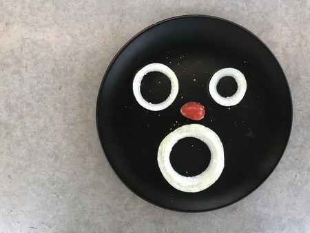 three pieces of onion and half of red grape tomato displayed on black plate looks like human face putted on gray cement bonded particle board with copy space on left side of frame, âplayfulâ conceptの写真素材