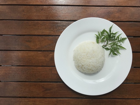 top view shot of plain Thai rice on white plate decorated with green leaf herb put on brown wooden table with copy space on right side of frameの写真素材
