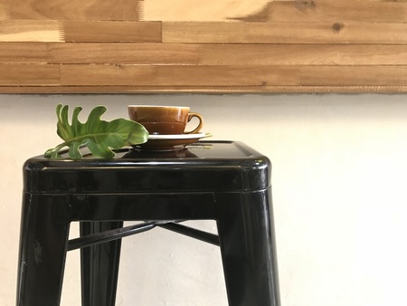 brown cup of hot coffee with bottom plate and green plant leaf on black metal bar stool with pieces...