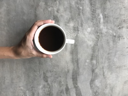 woman holding a white cup of hot black Americano coffee with her left hand on viva board or cement board table in coffee shop with copy space on right side of frameの写真素材